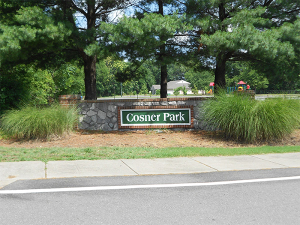 Virginia Custom Built Homes: Cosner Park sign surrounded by trees with a sidewalk and road in front of it